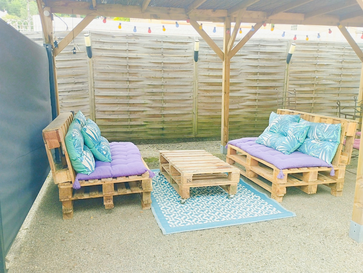 An outdoor patio area features two wooden pallet sofas adorned with vibrant cushions. A low wooden table sits at the center, complemented by a decorative area rug beneath. The space is partially shaded by a wooden structure, with string lights enhancing the setting.