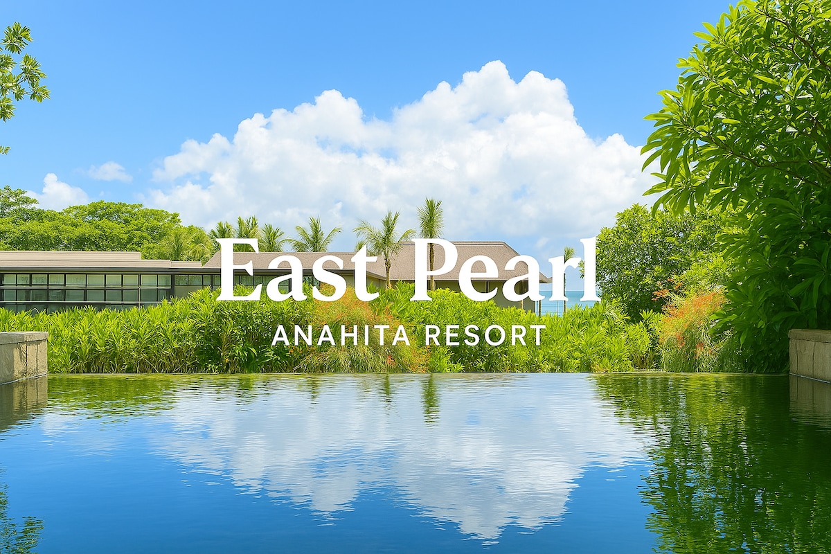 A tranquil view of the Anahita Resort is presented, featuring a reflecting pool that mirrors the lush greenery and blue sky. The surrounding tropical landscape enhances the serene atmosphere, creating a peaceful setting for relaxation.