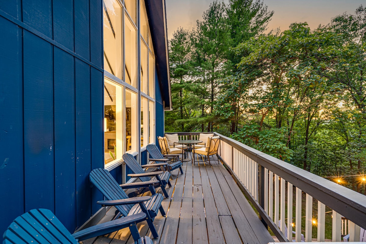 A spacious deck features several blue adirondack chairs arranged around a small table, providing a relaxed seating area. Large windows reflect the natural surroundings, with trees visible beyond the railing. Soft evening light enhances the inviting outdoor space.