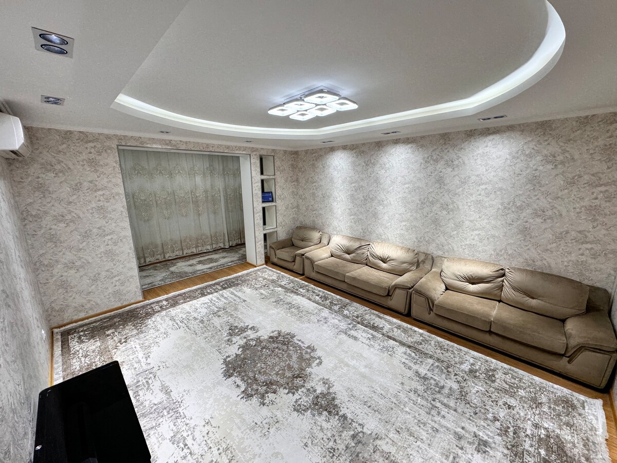 A spacious living area is furnished with a large, comfortable sectional sofa arranged around a light-colored area rug. Soft overhead lighting creates a warm ambiance, while a sliding door leads to a separate space, enhancing the room's functionality and openness.