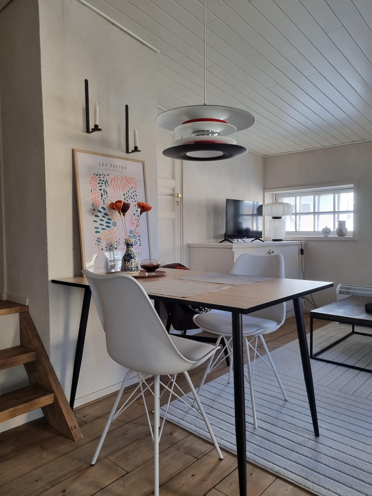 A dining area features a modern wooden table paired with white chairs. A colorful artwork adorns the wall above, accompanied by candles in sconces. A television is positioned against the wall, and natural light enters through multiple small windows.