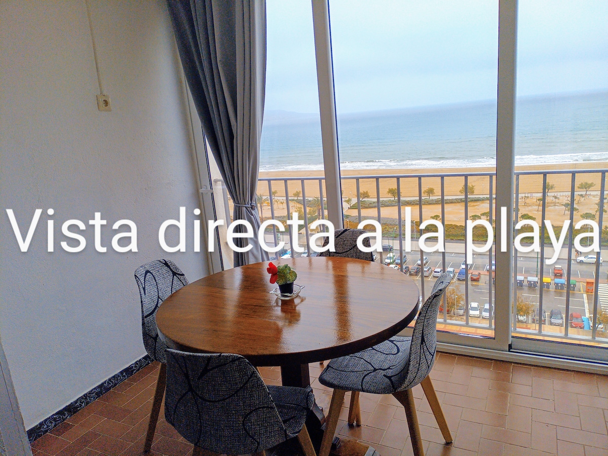 A dining area is shown, featuring a round table with four chairs. Large glass doors provide direct views of the beach and coastline. Natural light illuminates the space, enhancing the seaside atmosphere. Simple decor includes a small vase with flowers on the table.