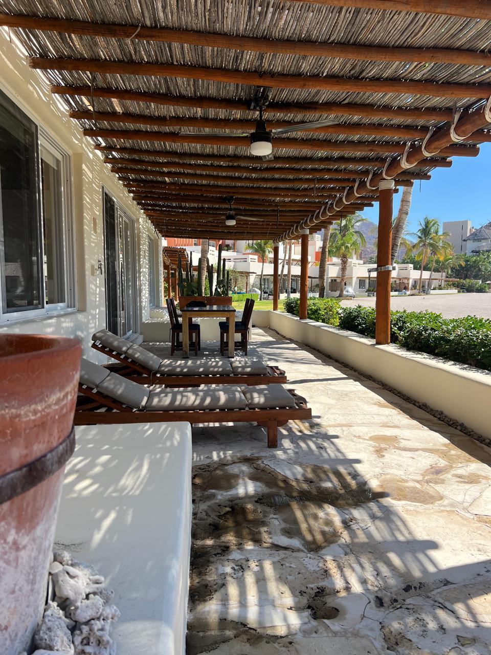 A shaded patio area features a long wooden table surrounded by chairs, perfect for outdoor dining. Lounge chairs are positioned for relaxation, and a potted plant sits nearby. Natural light filters through the overhead straw awning, creating a bright yet serene ambiance.