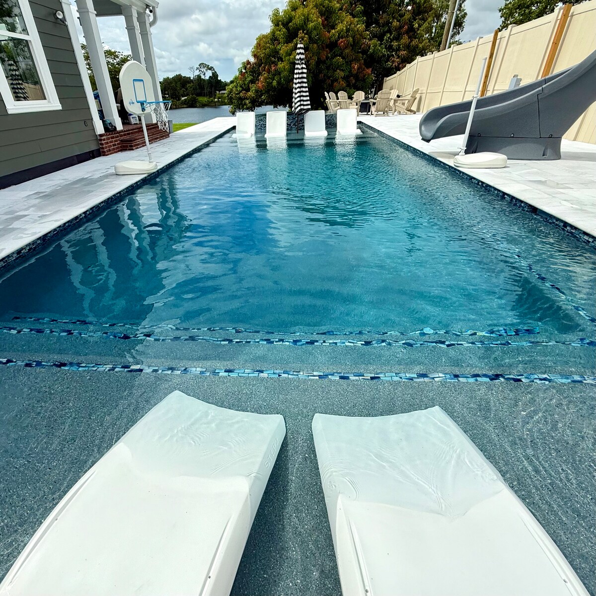 The image showcases a spacious swimming pool surrounded by a tiled deck. Two white lounge chairs are positioned at the edge, offering a view of the clear water. A slide and multiple seating areas are visible, enhancing the poolside experience.