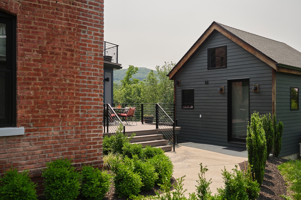 A newly renovated barn with a dark grey exterior is surrounded by greenery and features a small patio. The area is characterized by a mix of natural landscaping and modern architecture, set against a backdrop of rolling hills.