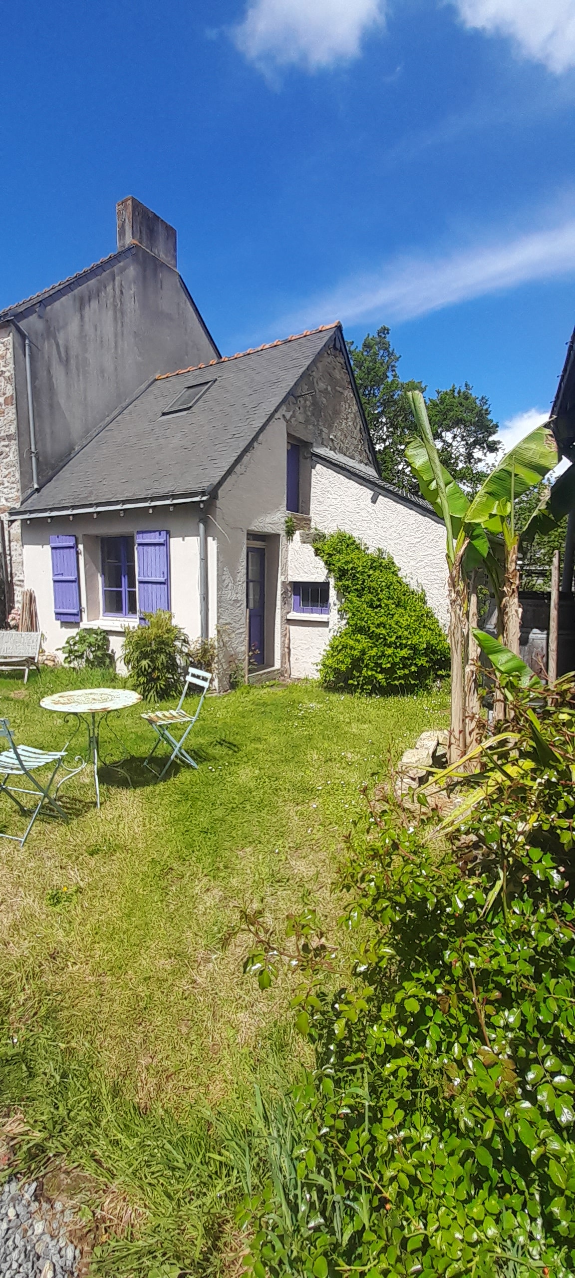 A charming stone house is surrounded by a lush garden, featuring vibrant green grass and a patio area with light green chairs. Lavender shutters adorn the windows, while tropical plants frame the entrance, adding to the inviting outdoor space under a clear blue sky.