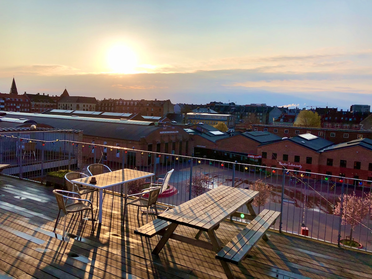 A rooftop terrace is shown with a picnic table and metal chairs, offering a view of the skyline and a setting sun. The wooden decking reflects warm light, and colorful decorations are suspended above the space, enhancing the outdoor atmosphere.