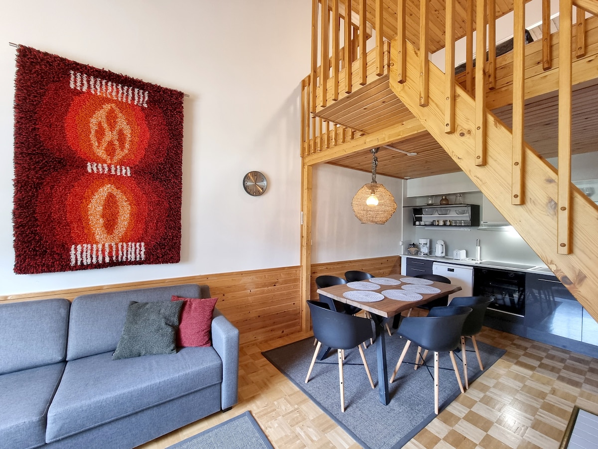 An open living and dining area features a grey sofa with colorful throw pillows. A round dining table with black chairs is positioned beneath a woven pendant light. The wooden staircase leads to the upper level, while a textured wall art piece adds visual interest.