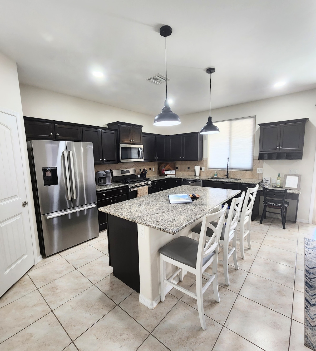 An open kitchen features dark cabinetry and granite countertops with modern appliances, including a stainless refrigerator and sleek range. A central island with seating for four is highlighted, while bright overhead lighting creates an inviting atmosphere. Large windows allow natural light to fill the space.