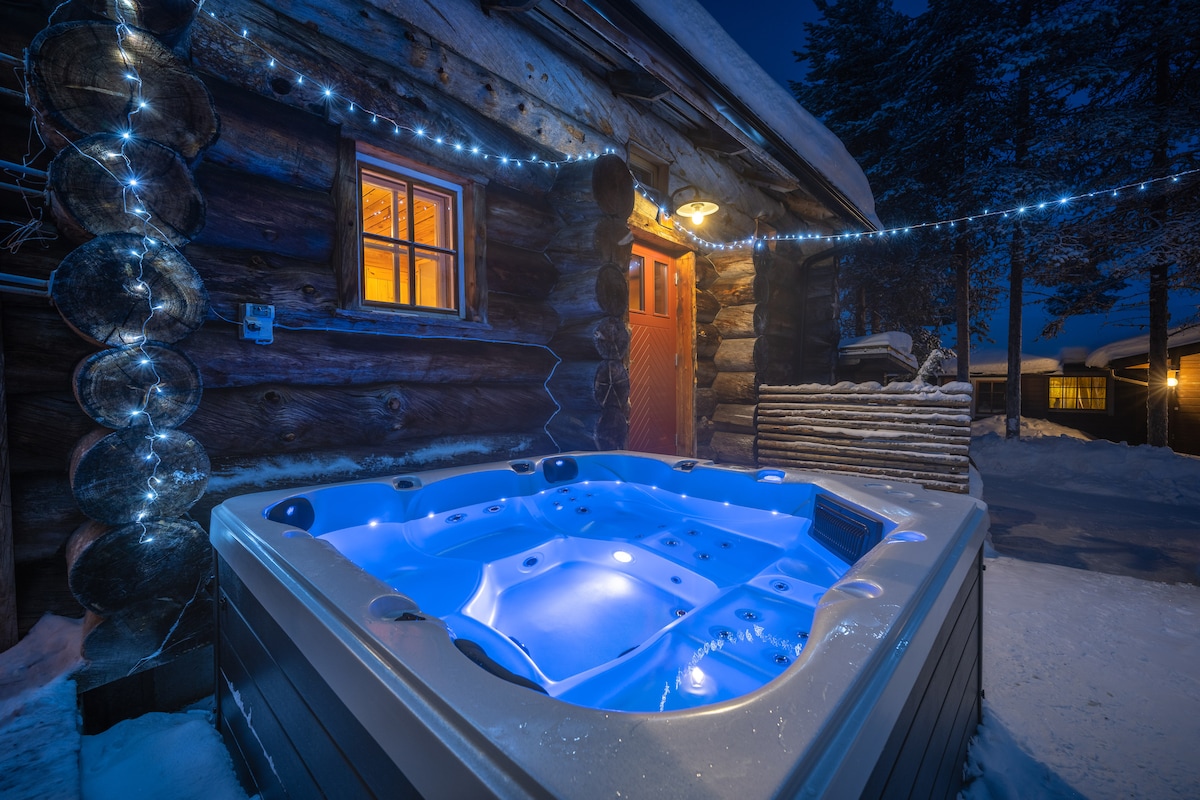 An outdoor hot tub is illuminated in soft blue light against a backdrop of snow-covered logs. The entrance to the cabin features a warm glow from a nearby window, complemented by a string of lights that add a festive touch to the wintery scene.