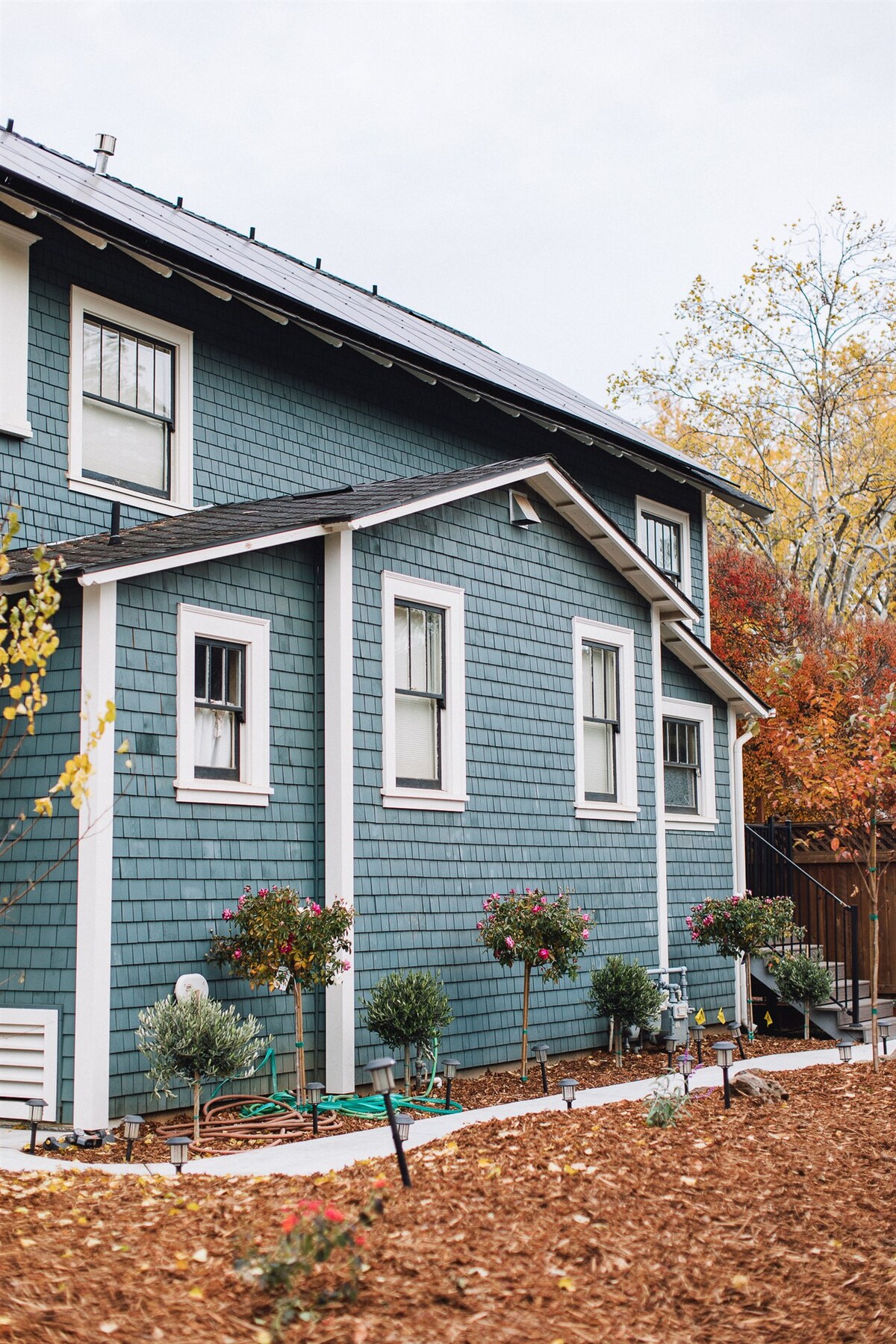 The exterior of a two-story blue cottage features textured shingles and solar panels. Window boxes contain vibrant flowers, and a pathway is flanked by small bushes. Surrounding trees depict autumn colors, enhancing the serene atmosphere of the property.