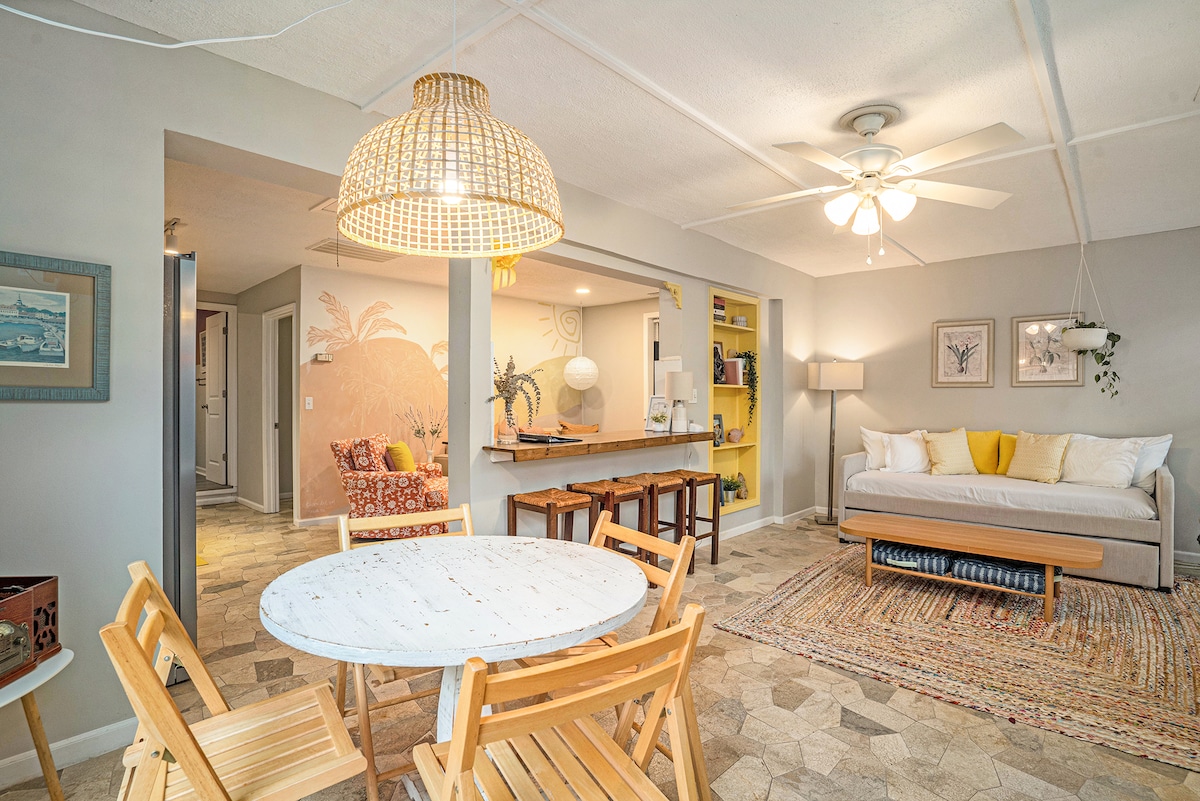 A bright living area features a round table with four wooden chairs. A daybed with decorative cushions is positioned nearby, complemented by a textured area rug. The space is illuminated by a large woven pendant light, enhancing the relaxed ambiance.