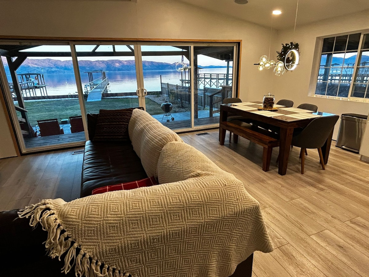 A cozy living area is framed by large glass sliding doors, offering expansive views of the lake. A spacious sectional sofa is draped with a textured throw, while a wooden dining table is positioned nearby. Natural light illuminates the room, enhancing the inviting atmosphere.