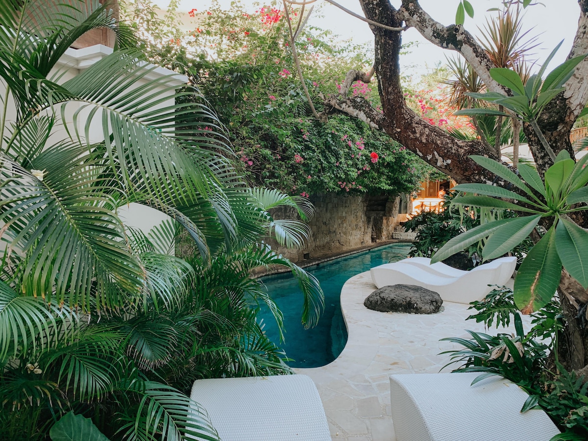 A serene outdoor pool area is surrounded by lush greenery and vibrant flowers. Smooth white lounge chairs are arranged beside the pool, which is bordered by natural stone. Tropical plants create a peaceful, secluded atmosphere, enhancing the sense of tranquility in the garden setting.