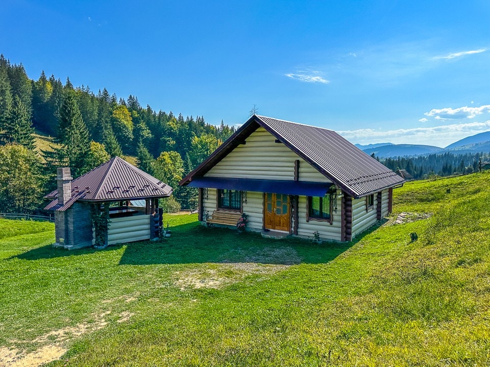 The exterior features a charming wooden house and a matching open gazebo, set against a backdrop of lush greenery and distant hills. Bright blue sky and sunlight illuminate the structure, highlighting the natural surroundings that enhance the peaceful ambiance.
