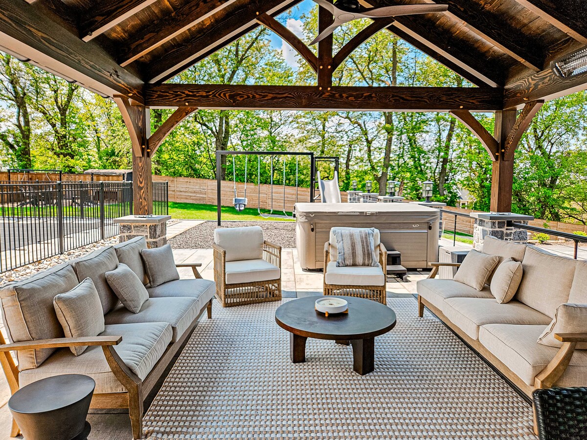 An outdoor seating area is covered by a wooden pergola, featuring comfortable beige sofas arranged around a dark circular table. A hot tub is positioned in the background, surrounded by greenery. The flooring is stone, adding to the inviting space.