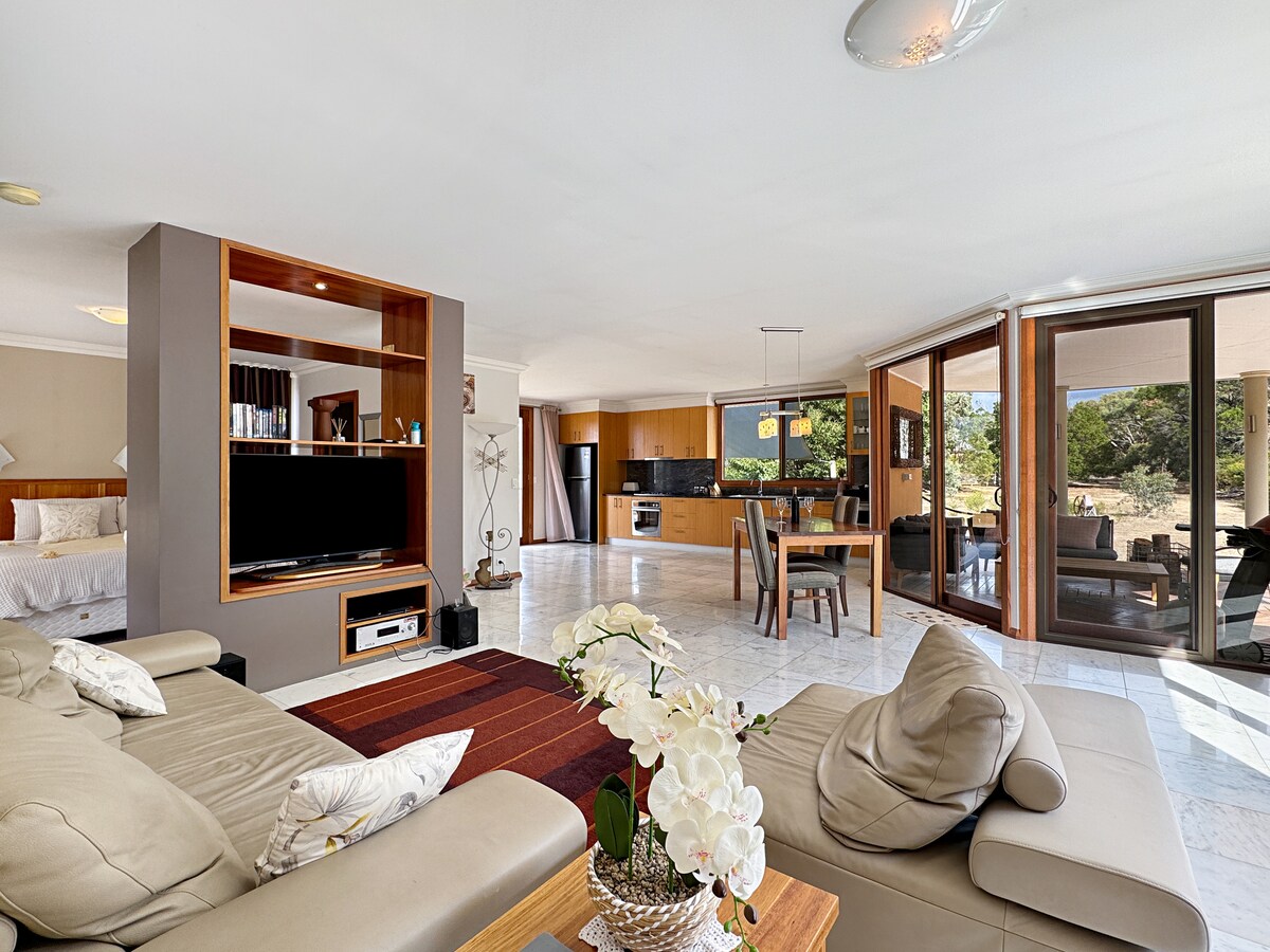 An open-plan living area features a comfortable lounge setup with a soft couch and elegant decor. The kitchen and dining space are visible in the background, with large glass doors offering a view of the surrounding nature. Natural light illuminates the warm-toned furnishings.