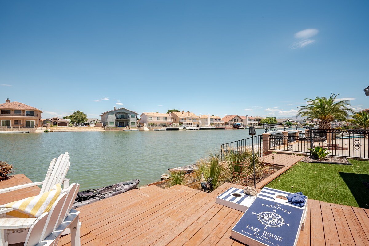 A spacious back deck features wooden decking with lounge chairs facing a serene lake view. Lush greenery and decorative elements are included, while the water is framed by neighboring homes. The scene is illuminated by clear skies and natural light.