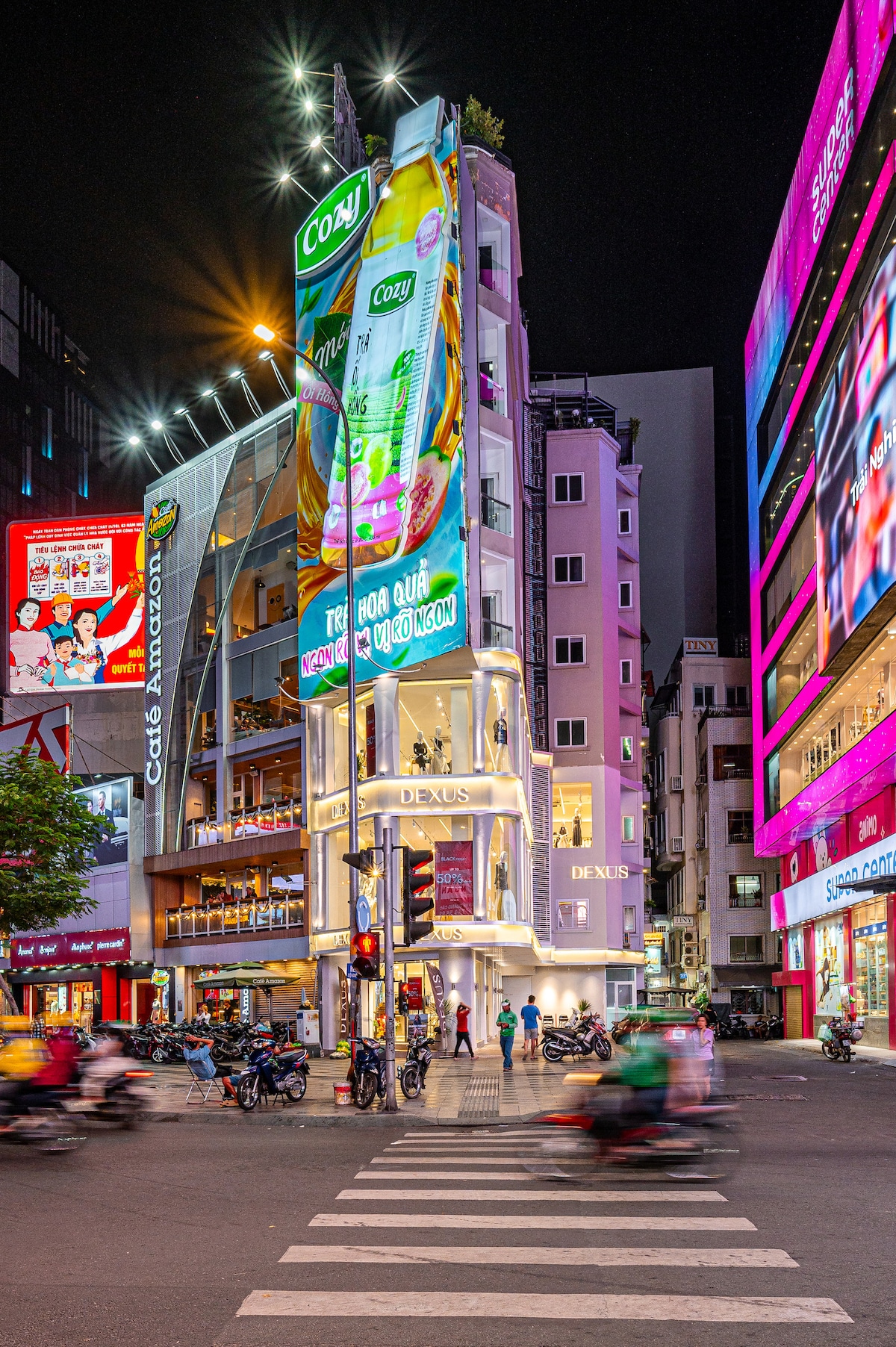 A vibrant urban scene captured at night, showcasing a multi-story building adorned with colorful advertising displays. Bright lights illuminate the surrounding area, while street traffic flows past, highlighting the lively atmosphere of downtown Saigon.