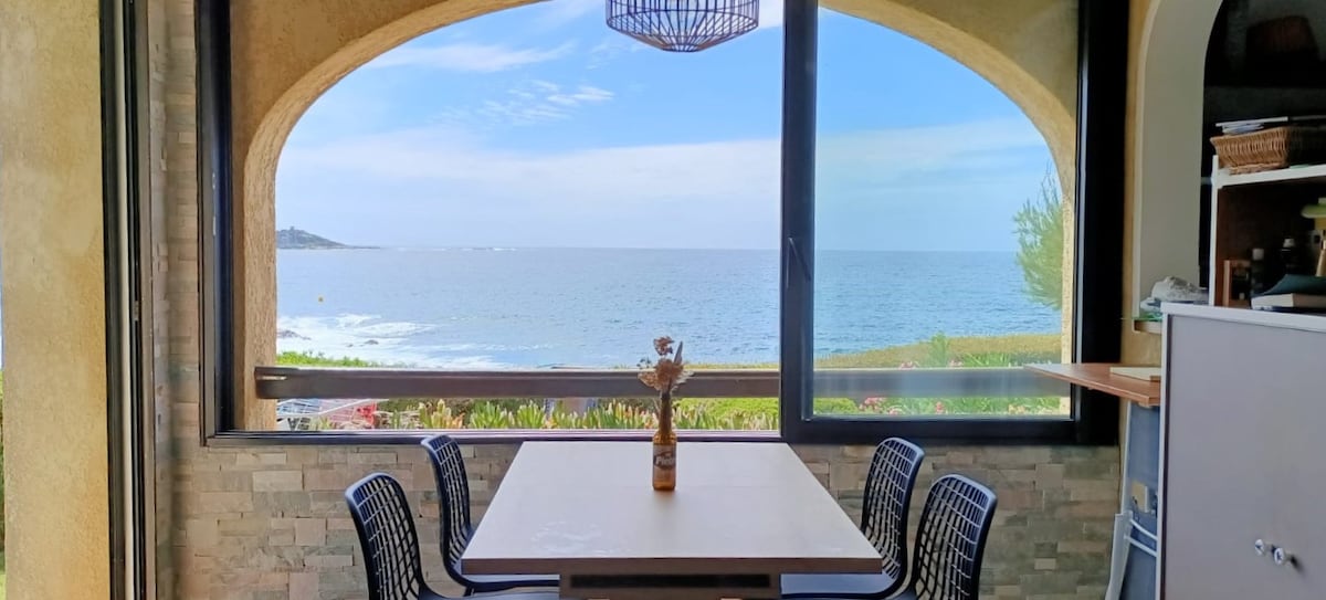 A dining area is framed by a large arched window that offers a direct view of the sea. A simple table is set with a vase at its center, surrounded by four black chairs. Natural light fills the space, enhancing the coastal atmosphere.