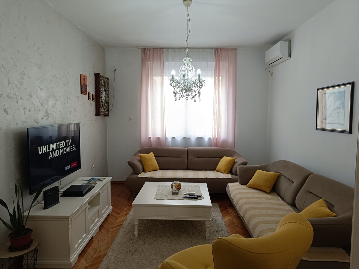 A living room features a combination of comfortable beige and yellow seating arranged around a light coffee table. A flat-screen television is positioned on a white media unit. Natural light is diffused through gently sheer curtains, complemented by a decorative chandelier overhead.