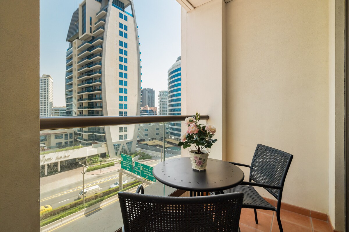 A balcony area features a round table and two black chairs, offering a view of the surrounding buildings. A potted plant adds a touch of greenery, while the urban landscape stretches beyond, highlighting the vibrant atmosphere of the neighborhood.
