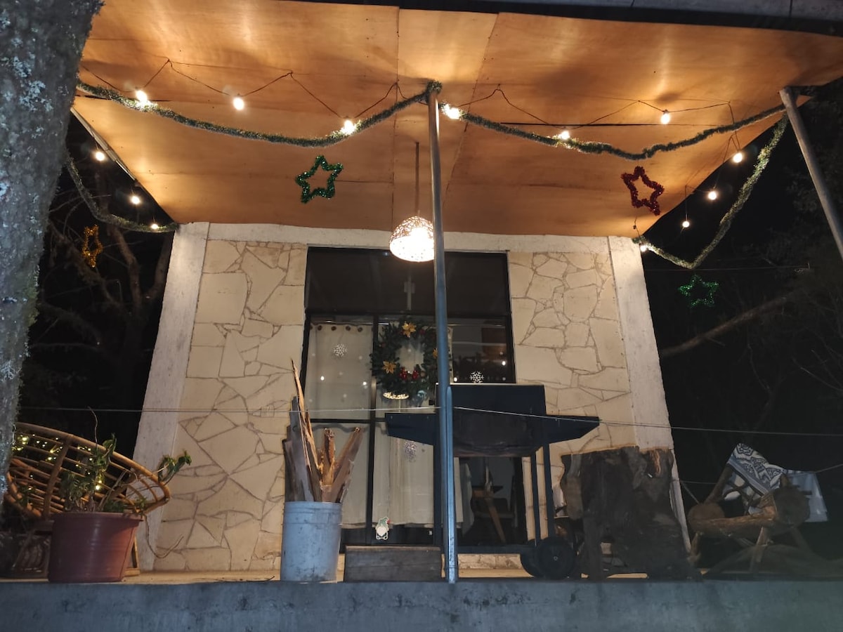 A covered outdoor area features a wooden table and chairs, surrounded by potted plants. Decorative lights and stars adorn the ceiling, creating a festive ambiance. A view through the large glass doors highlights a cozy interior space.