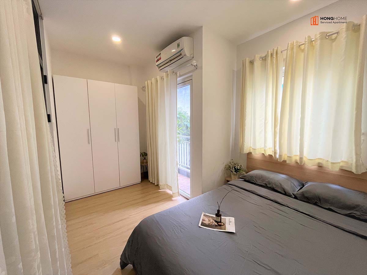 A well-lit bedroom features a spacious bed with gray linens positioned against a wooden wall. Nearby, white curtains frame a window leading to a balcony. An organized white wardrobe stands beside the bed, enhancing the room's modern design.