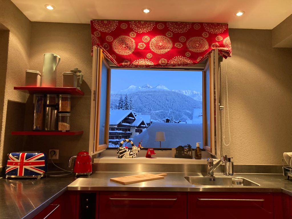 The kitchen features a modern counter with a stainless steel sink and ample storage. A window offers a view of snow-covered mountains and chalets. The colorful window treatment complements the red shelves holding kitchenware, while small appliances add functionality.