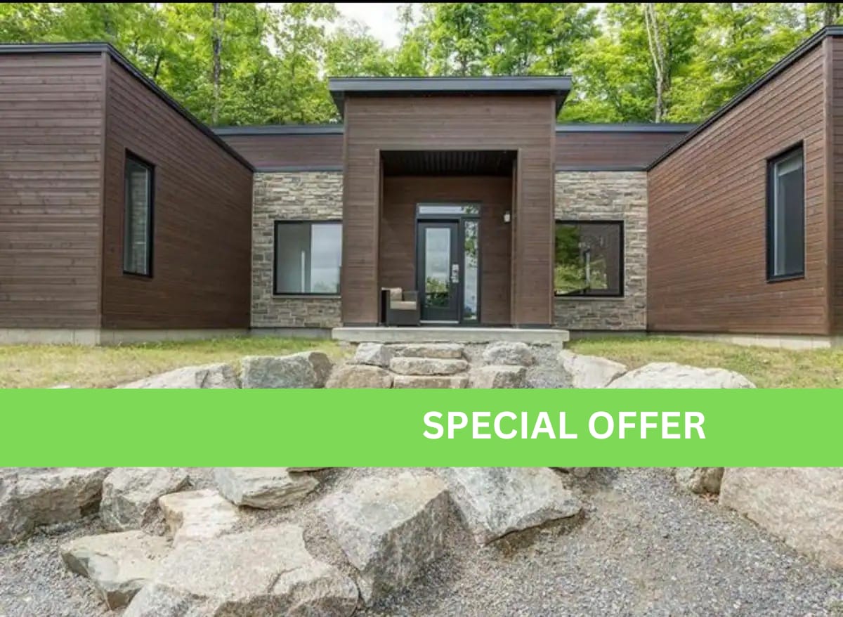 A modern chalet exterior is framed by a combination of wood and stone cladding. Large windows provide a view into the interior. A pathway of stone leads to the front door, while natural landscaping features rocks and grassy areas.