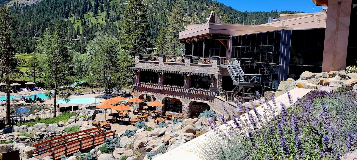 An outdoor space features a series of pathways leading among landscaped areas. Patio tables with orange umbrellas are arranged near a clear pool, bordered by stone structures. The backdrop showcases a building with large windows and a wooden balcony, set against a mountainous landscape.