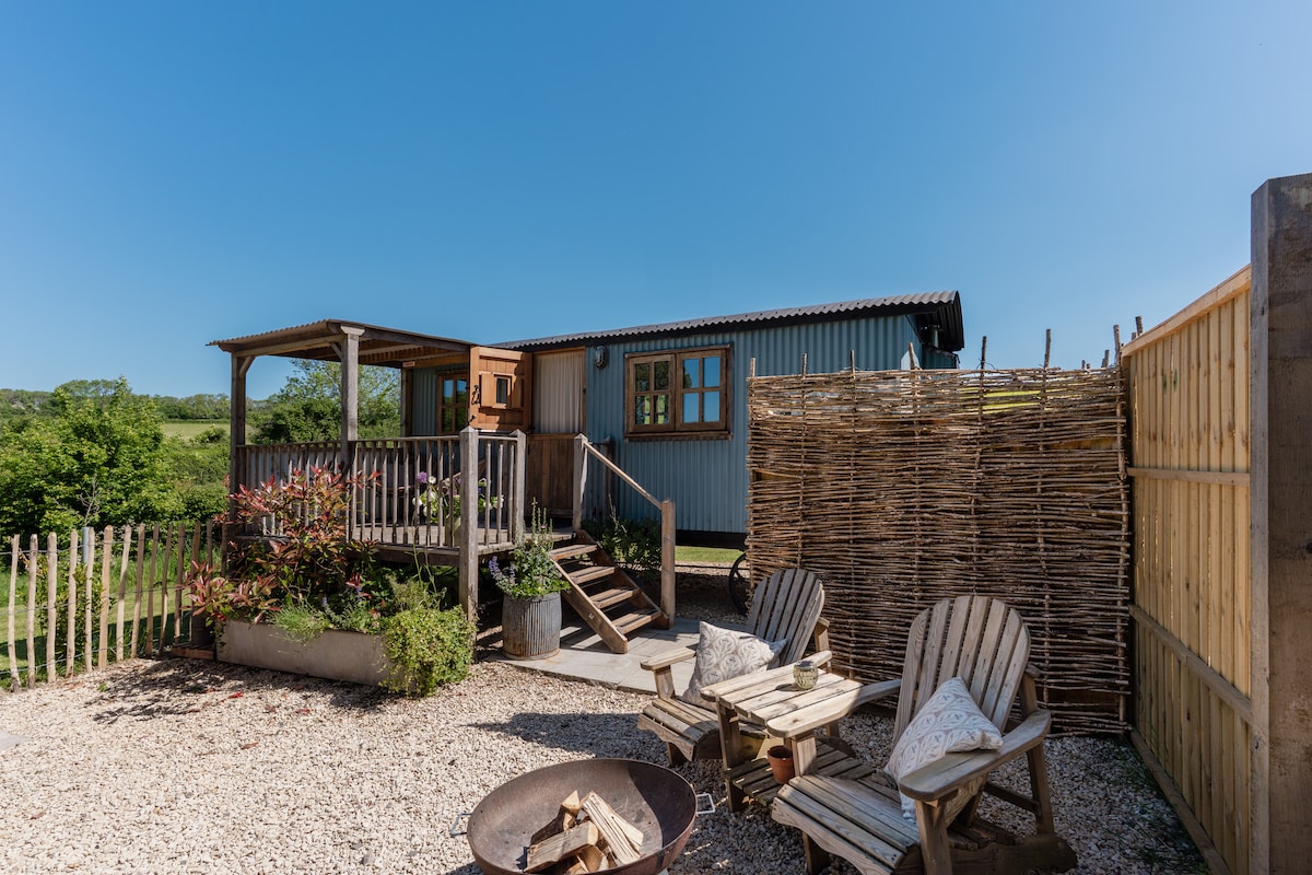 A charming outdoor area displays a wooden deck leading to the shepherd's hut, surrounded by lush greenery. Two rustic chairs are positioned around a fire pit, inviting relaxation. A woven fence and planter add natural elements to the tranquil setting under a clear blue sky.