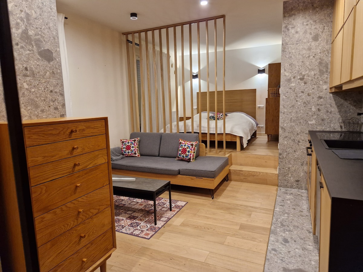 A stylish and open studio layout presents a cozy seating area with a gray sofa and decorative cushions. A small coffee table is positioned next to a patterned rug, while a well-defined sleeping space features a spacious bed framed by wooden partitions, ensuring privacy and comfort.