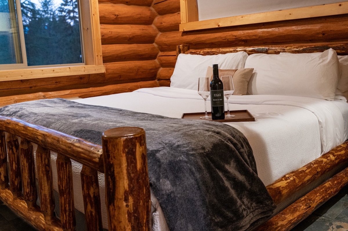 A spacious bed is framed by rustic wooden logs, creating a cozy sleeping area. A tray holds a bottle of wine and two glasses, positioned atop a soft white blanket. Natural light enters through a window, enhancing the cabin's welcoming atmosphere.