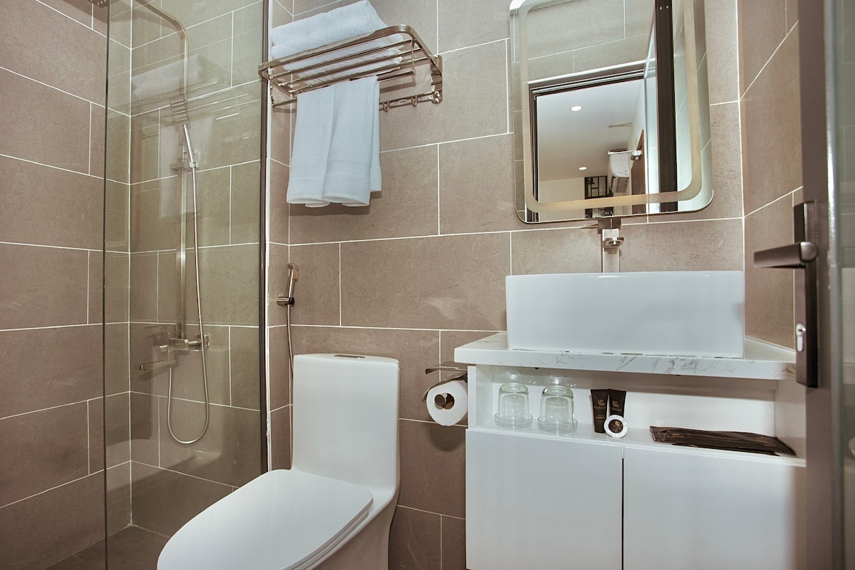 A modern bathroom features a glass-enclosed shower with sleek fixtures. A minimalist white sink is complemented by a wall-mounted mirror. Fresh towels are neatly arranged on a rack, and essential toiletries are stored underneath the sink, providing practicality and convenience.
