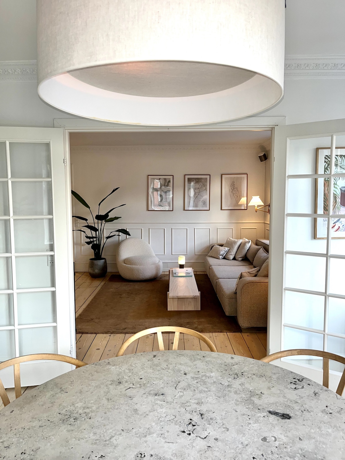 A light-filled living space is visible through open French doors, featuring a comfortable gray sofa and a modern coffee table on a neutral area rug. A large pendant light hangs above a circular dining table with wooden chairs, enhancing the airy atmosphere.