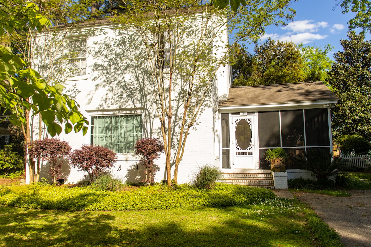 The two-story country home features a front porch with a screen, framed by lush greenery and flowering plants. Bright natural light is evident, highlighting the white brick façade and large windows. A peaceful yard showcases well-maintained landscaping, creating a serene outdoor environment.