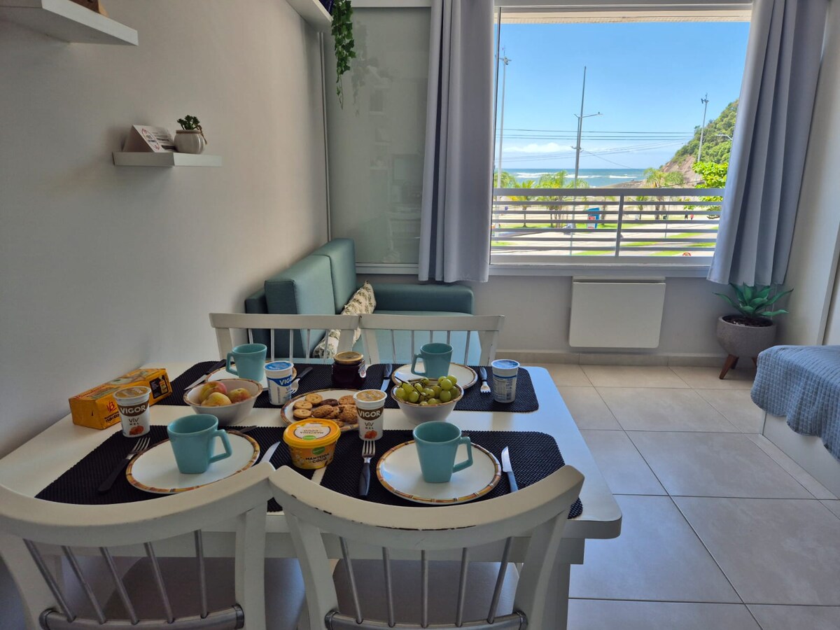 A bright dining area features a white table set with plates, cups, and an assortment of snacks. A cozy sofa is positioned beside a large window, showcasing a view of the ocean and surrounding landscape. Soft curtains frame the window, adding to the inviting atmosphere.