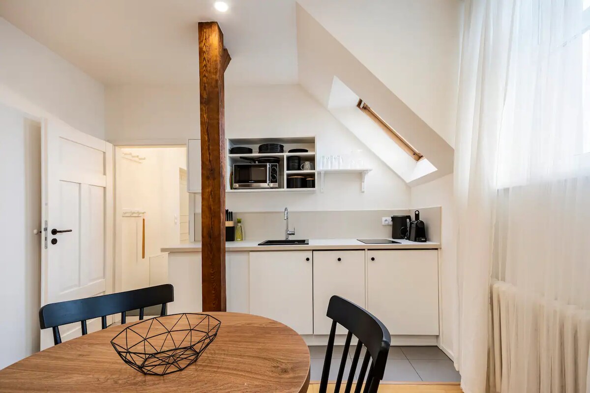 A compact kitchen area features modern appliances, including a microwave and coffee maker, set against a backdrop of light cabinetry. A round wooden table with black chairs provides a dining space, while natural light filters through a nearby window.