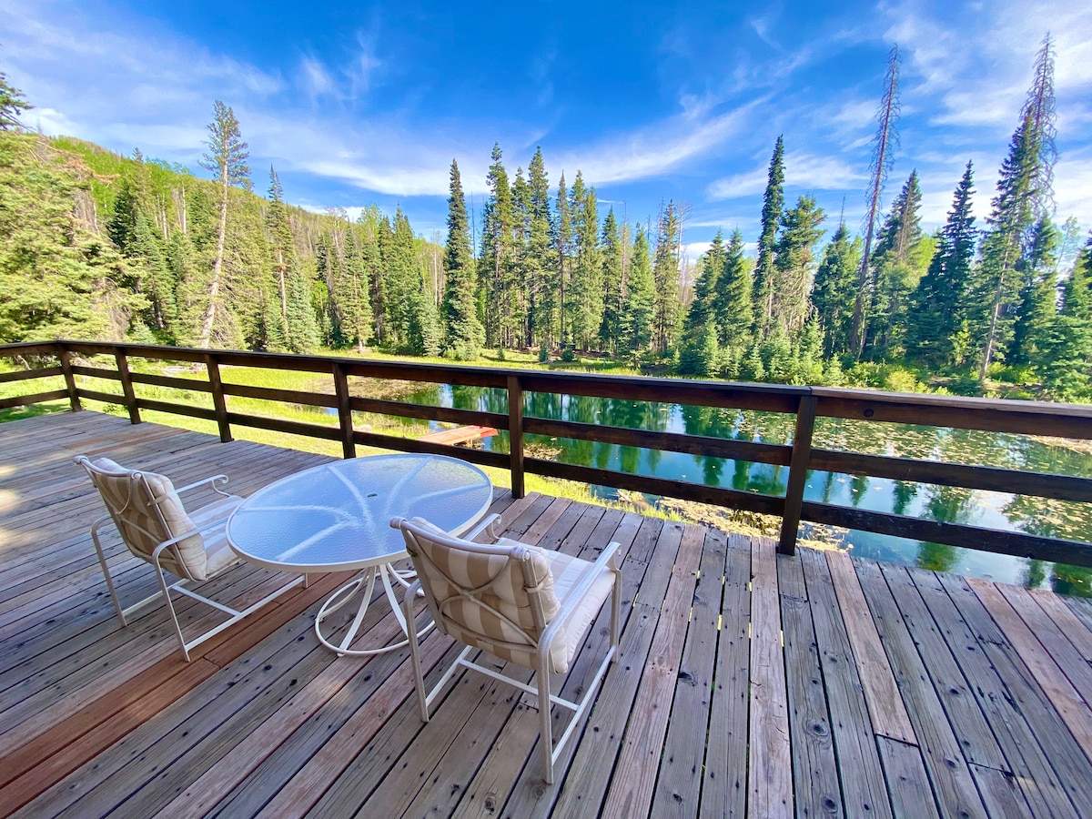 A covered deck features a round glass table surrounded by two light-colored chairs. The scene overlooks a serene private pond, framed by lush trees and greenery under a bright, blue sky.