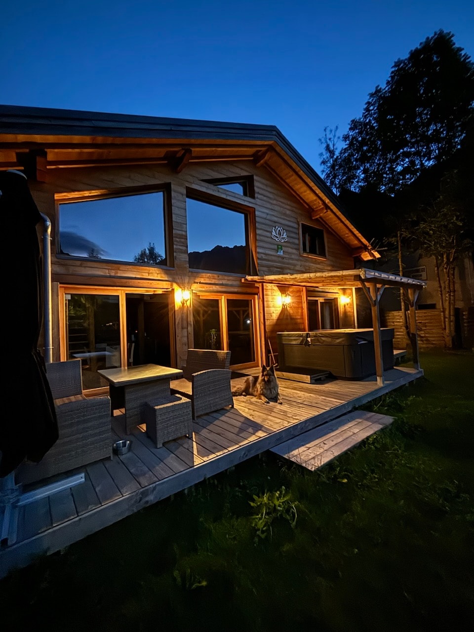 The exterior of a chalet is captured at night, featuring large glass windows that reflect warm light. A wooden deck is furnished with a small table and chairs, while a hot tub is positioned nearby, surrounded by lush greenery.