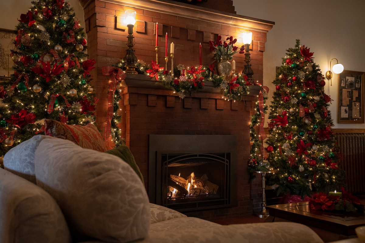 A beautifully decorated living room features two festive Christmas trees adorned with ribbons and ornaments. The red brick fireplace, set with a gas insert, is surrounded by seasonal decorations, while soft lighting creates a cozy ambiance. A plush sofa provides a comfortable seating area.