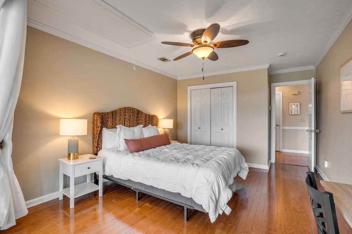 A spacious bedroom is furnished with a plush bed featuring a decorative headboard. Two bedside lamps offer soft lighting, and a wooden desk is positioned nearby. Light wood flooring enhances the room's warm atmosphere, with a ceiling fan providing airflow.