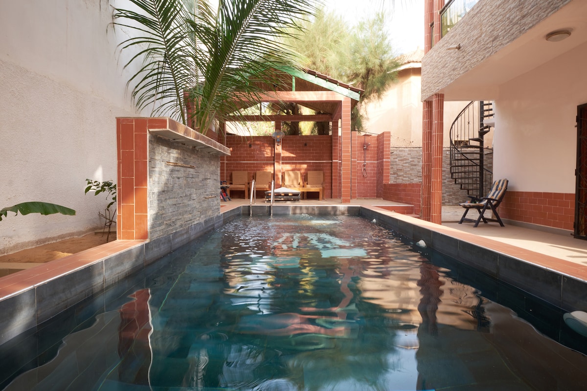 A private swimming pool is featured, surrounded by lush greenery and terracotta tiles. A natural stone wall provides a visual focal point. The area is complemented by a covered space containing seating for relaxation and enjoyment.