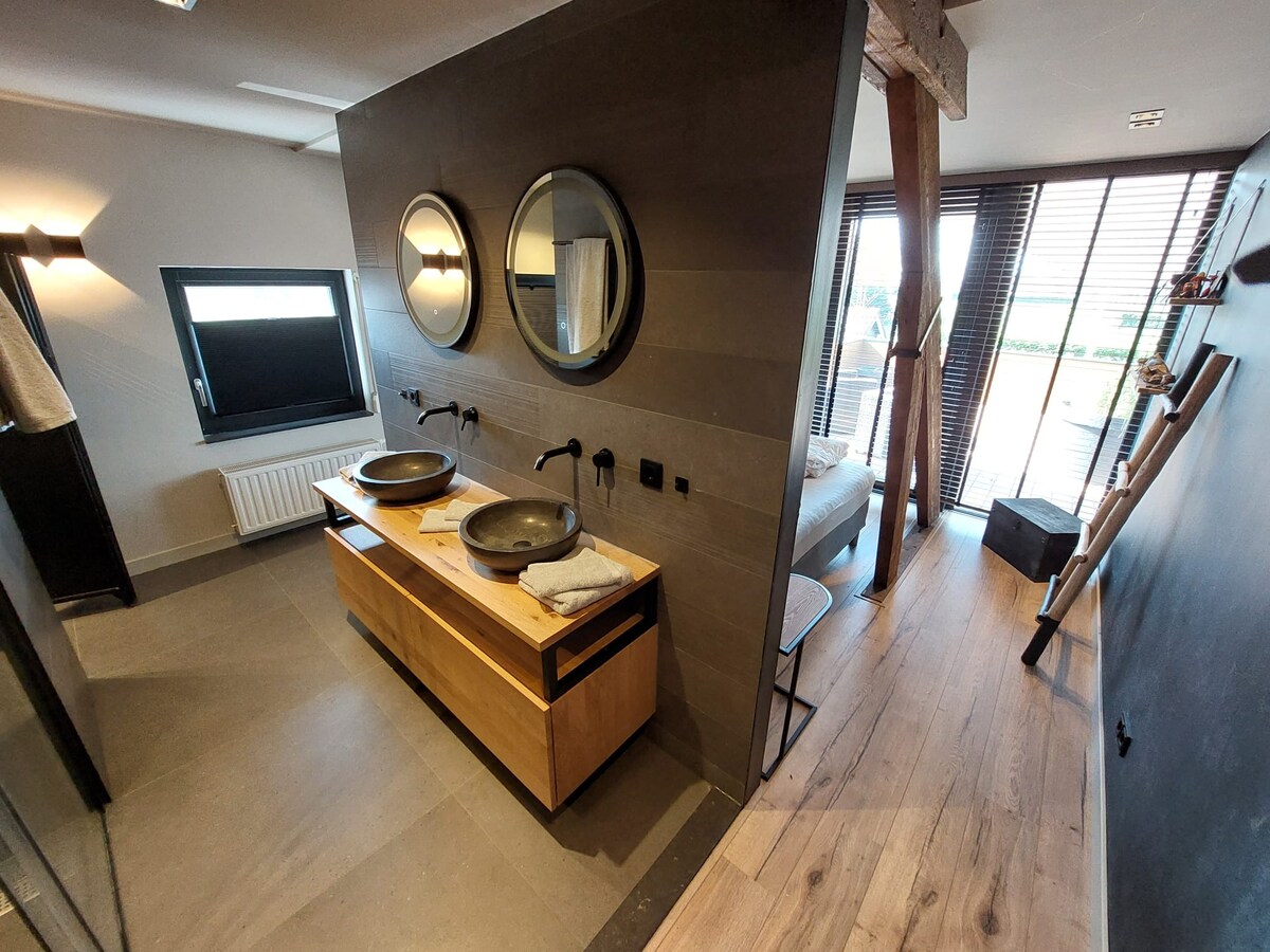 A modern bathroom features dual circular mirrors above stylish sinks. The countertop is crafted from natural wood, paired with dark faucet fixtures. Light streams in through window coverings, creating a serene atmosphere. The adjoining area includes a glimpse of a cozy sleeping space beyond.