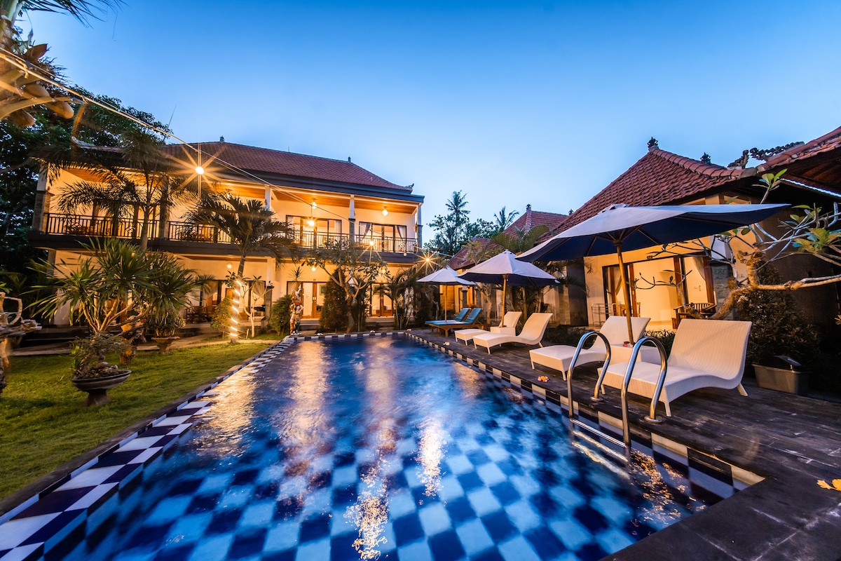 An outdoor swimming pool is bordered by loungers and umbrellas, surrounded by lush greenery. Two structures are visible in the background, featuring traditional Balinese architecture. The pool's water reflects the evening sky, creating a serene environment enhanced by the warm lights illuminating the area.
