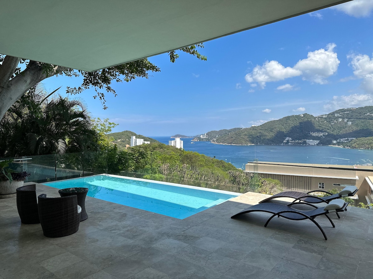 A spacious terrace features a private pool surrounded by glass railing, offering a view of the bay and distant hills. Two lounge chairs and two woven chairs are positioned nearby, contributing to a relaxing outdoor atmosphere.