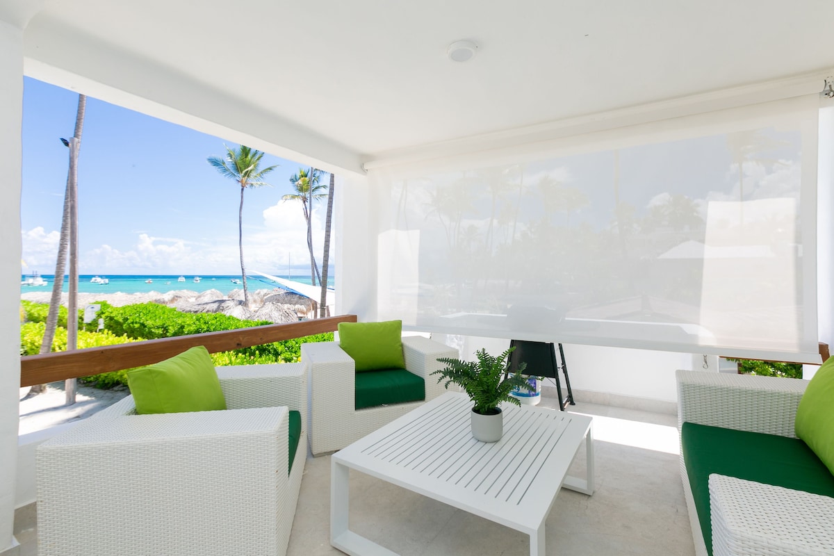 A shaded seating area showcases a white wicker table surrounded by four matching chairs, accented by bright green cushions. The view through large windows features a vibrant beach scene, with palm trees and blue ocean waters visible in the distance.