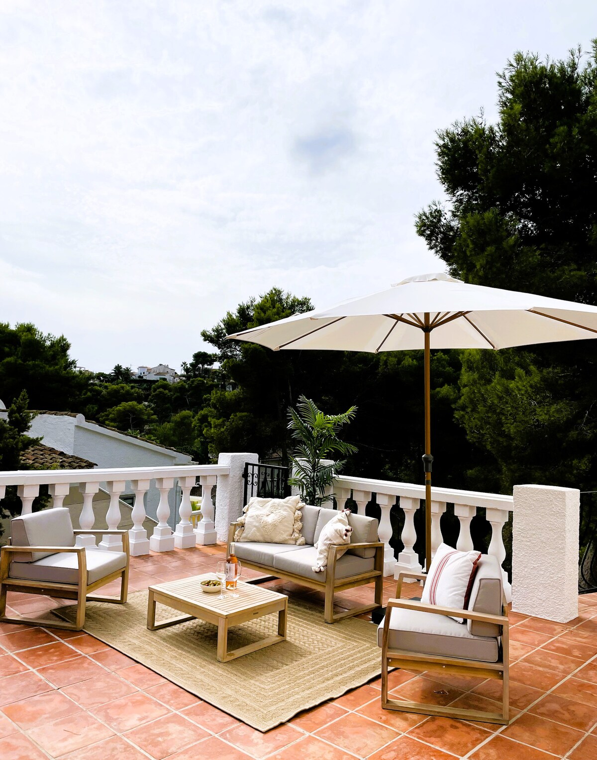 A terrace area features comfortable seating arrangements under a spacious umbrella. A light-colored rug is placed beneath a small table, providing a cozy spot for relaxation. The surrounding greenery enhances the peaceful ambiance, while decorative cushions add subtle detail.
