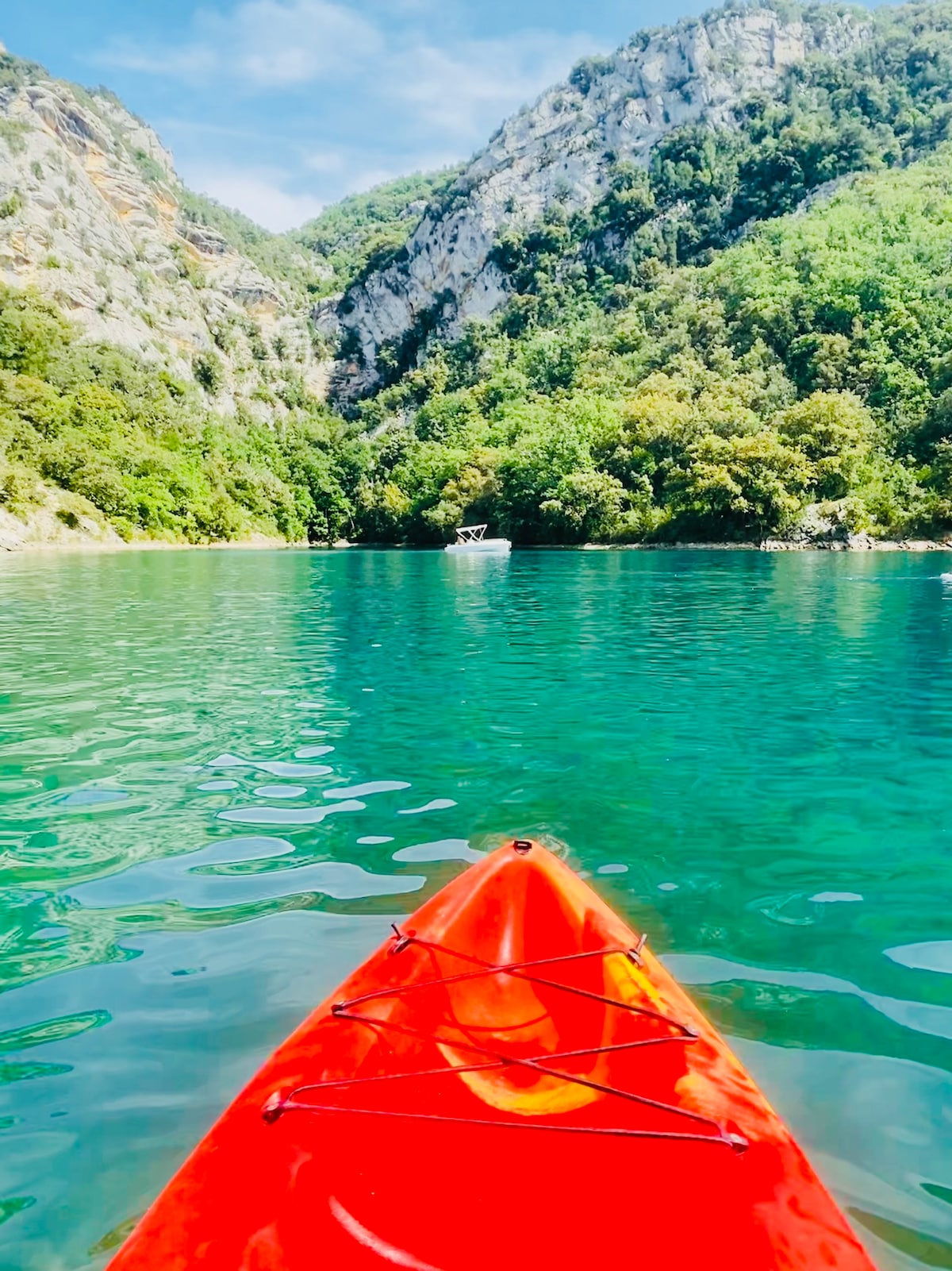 An orange kayak is positioned in tranquil waters, reflecting vibrant shades of blue and green. Lush greenery covers the surrounding cliffs, creating a natural backdrop. The serene atmosphere suggests an ideal setting for water activities and relaxation near the lake.